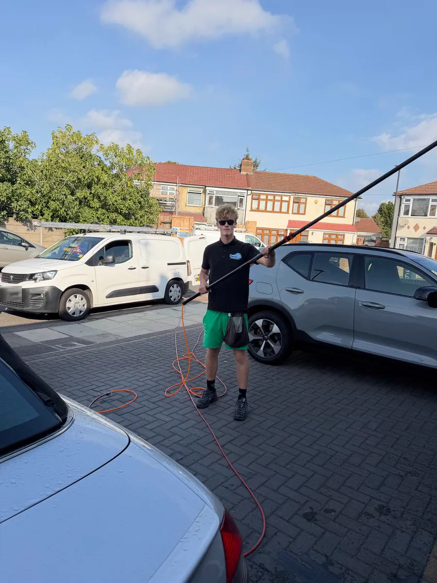 Teo's son in sunglasses and a Teo Exterior Cleaning polo holding a water-fed pole beside his van, with the hose on the paving around him
